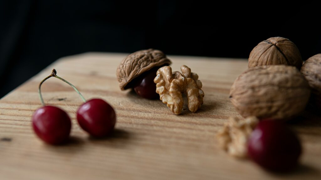 The image is a close-up photograph of a rustic wooden surface featuring a few walnuts in their shells and a walnut kernel, along with three cherries. One walnut is open, showing the edible part inside. The setting has a natural, subdued lighting that highlights the textures of the walnuts and the glossy surface of the cherries. The composition is simple yet elegant, emphasizing the natural beauty and texture of these fresh ingredients. The dark background enhances the rich colors and organic shapes of the walnuts and cherries, making this a visually appealing scene.